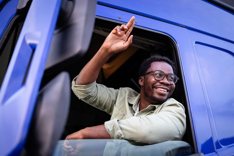 A smiling fleet driver prepares for delivery while waving from the cab of a semi-truck.