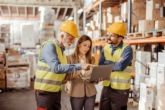 A business owner and managers with hard hats review data from a fuel card on a computer while standing in a warehouse.