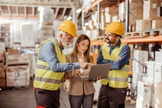 A business owner and managers with hard hats review data from a fuel card on a computer while standing in a warehouse.
