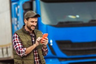 A happy truck driver uses his fleet fuel card app on his phone standing outside his rig.