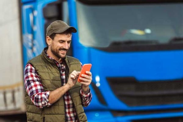 A happy truck driver uses his fleet fuel card app on his phone standing outside his rig.