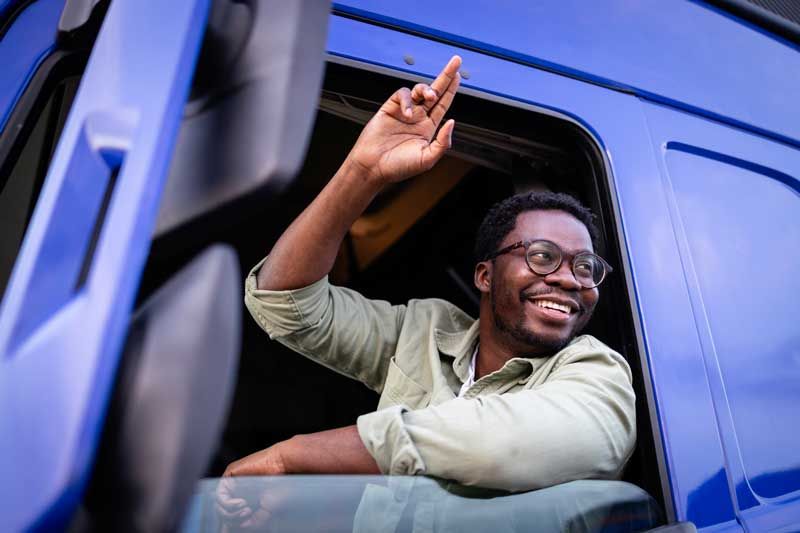 A smiling fleet driver prepares for delivery while waving from the cab of a semi-truck.